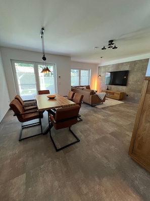 A modern living room with a dining area, a flat-screen TV mounted on a textured wall, and a ceiling fan. - Holiday Home 'Ferienhaus Am Nationalpark' with Lake View, Wi-Fi and Air Conditioning (Klausdorf)