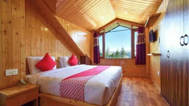 Un dormitorio de cabaña de madera con techo inclinado, una cama con almohadas rojas, una ventana con cortinas y piso de madera.