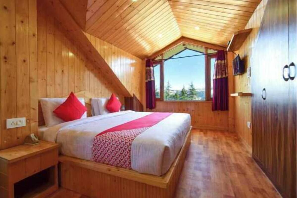 Une chambre dans une cabane en bois, avec un plafond en pente, un lit avec des oreillers rouges, une fenêtre avec des rideaux et un plancher en bois.