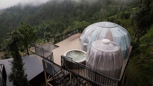 A transparent geodesic dome structure with a hot tub and a seating area, surrounded by a wooden deck and metal railings, set against a mountainous backdrop. - Castle Glamp by Auroras Resorts (Kosiyakutoli)