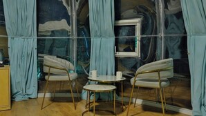 A room with a wooden floor, two white chairs with gold legs, a small round table, and a window with a view of a blue sky and clouds. - Castle Glamp by Auroras Resorts (Kosiyakutoli)