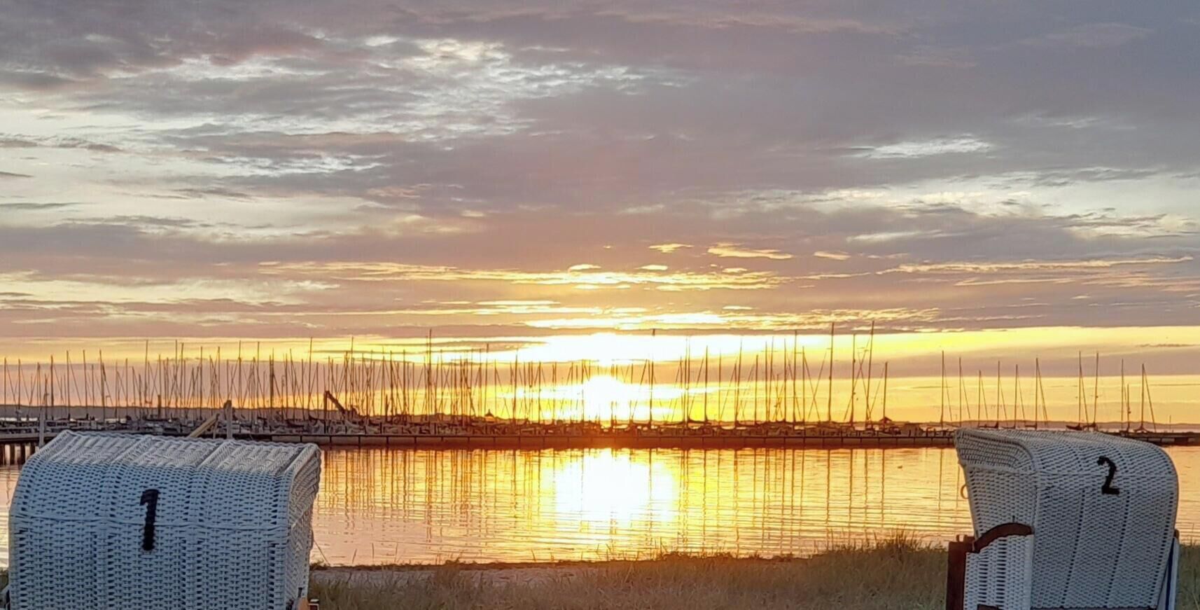 Ein Sonnenuntergang über einem Yachthafen mit Segelbooten, zwei Korbstühle mit den Aufschriften '1' und '2' sowie eine Holzsteganlage.