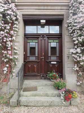 A double wooden door with a number plaque, a small step with a metal handrail, and a flower arrangement at the entrance.