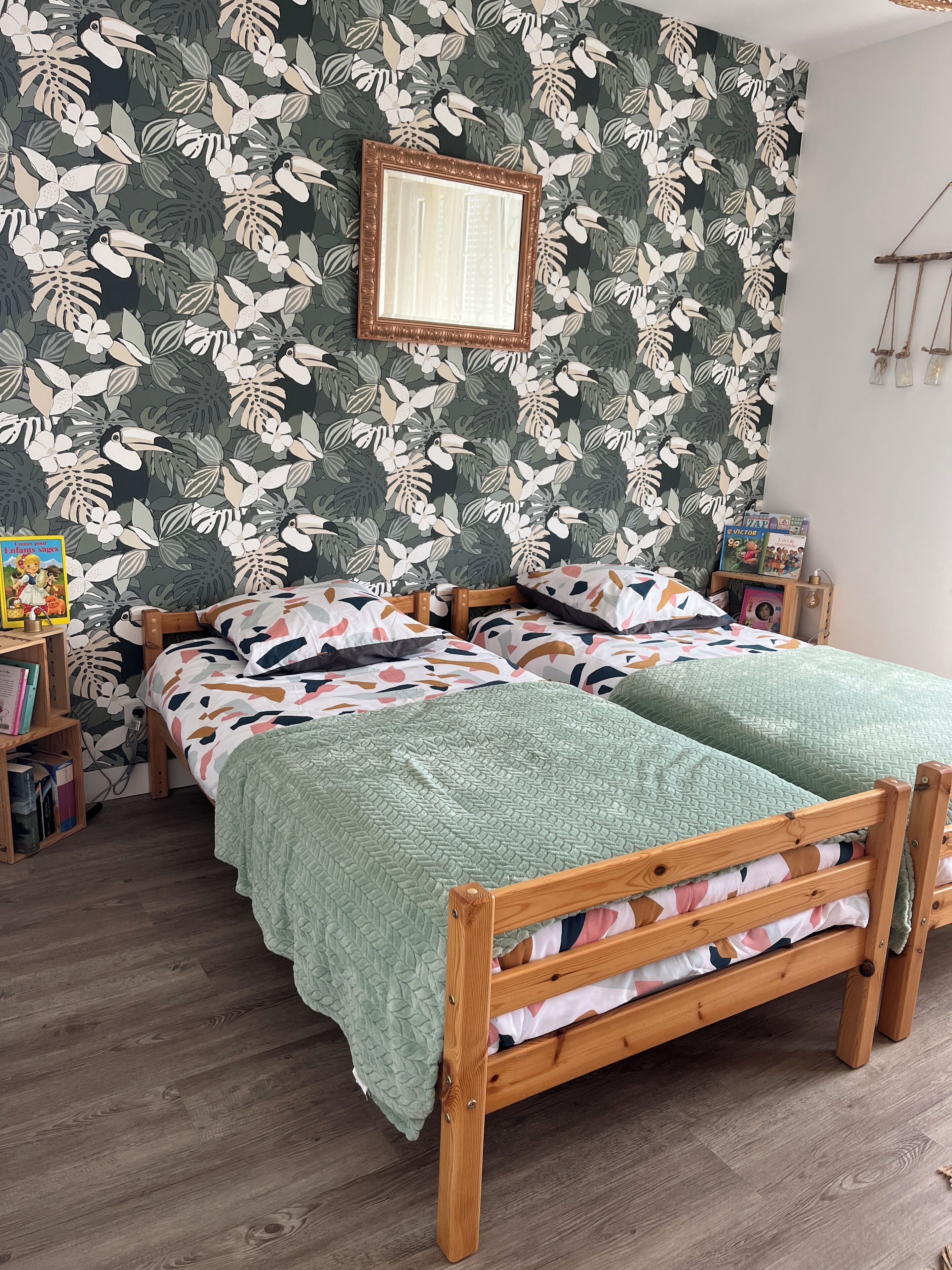 Un dormitorio con cama de madera, ropa de cama verde, una estantería y una pared con diseño de hojas tropicales.