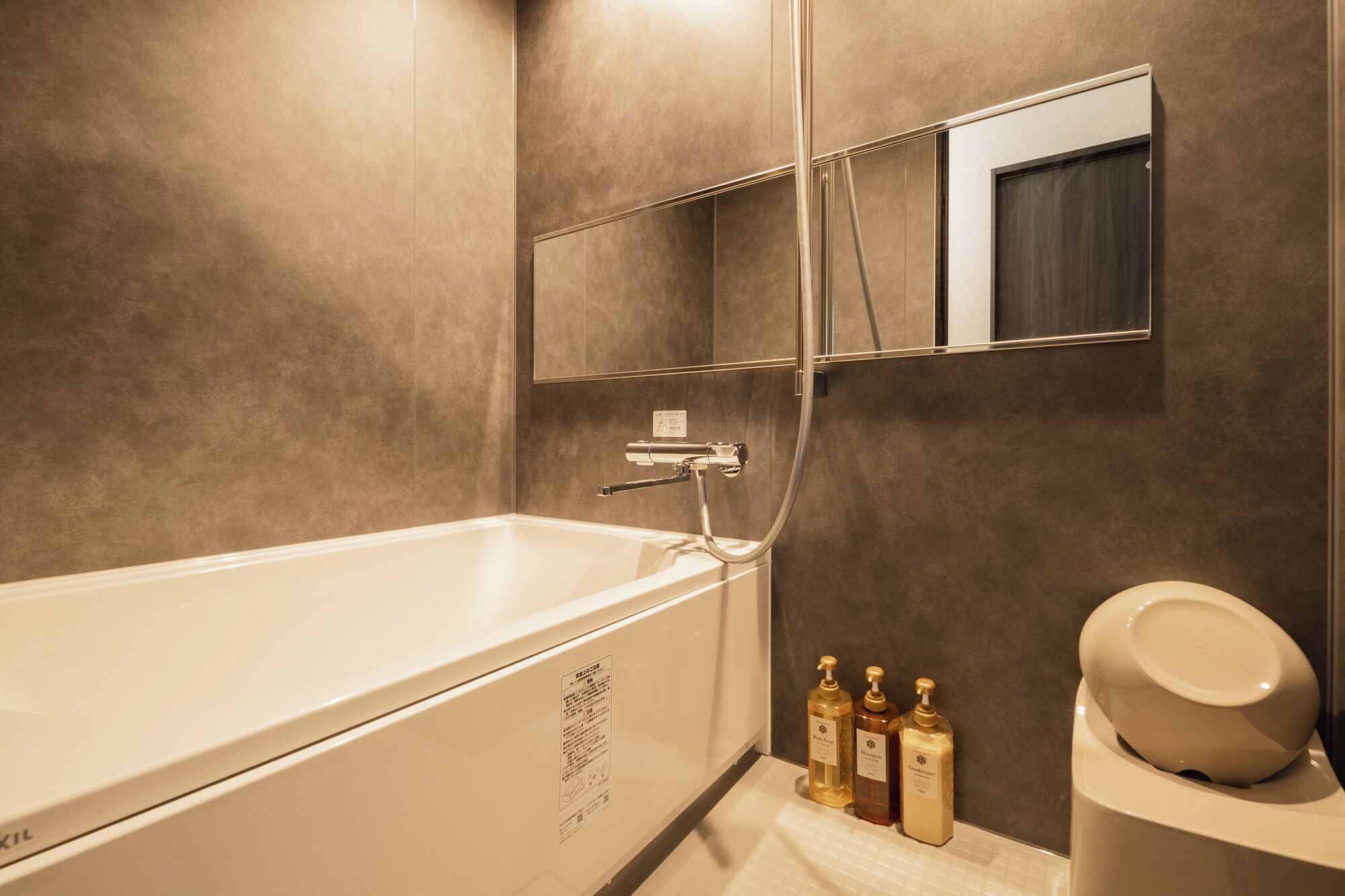 A modern bathroom with a bathtub, a toilet, and a mirror. There are three bottles of toiletries on the counter.