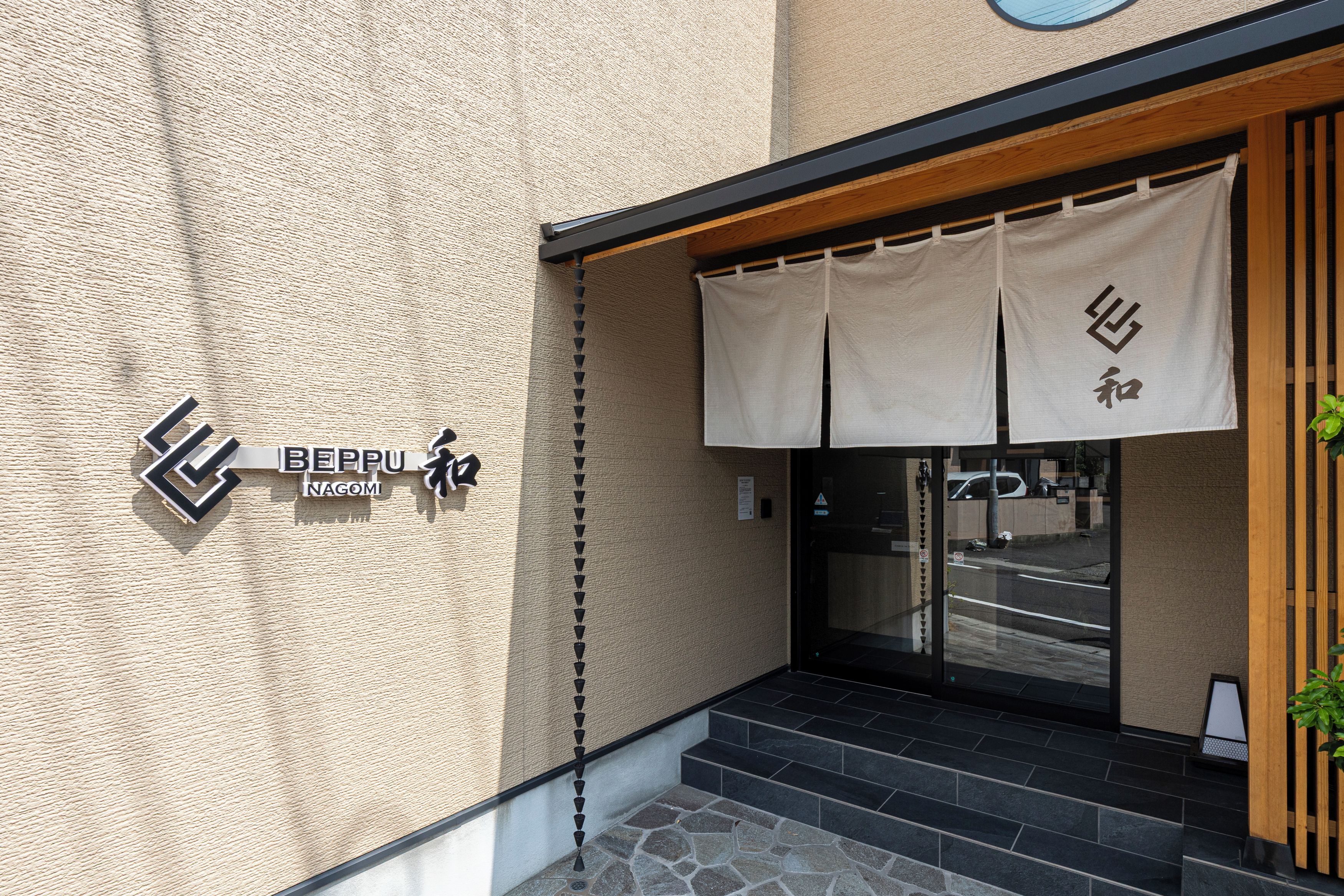 A building entrance with a textured wall, a sign reading 'BEPPU NAGOMI', and a wooden door with a curtain.