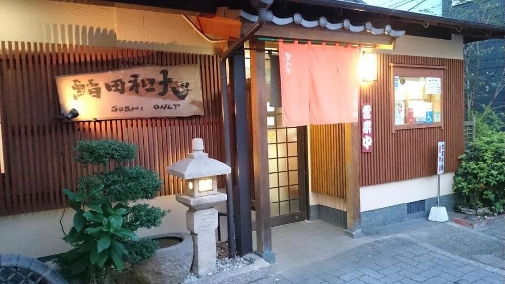Un restaurant de sushis avec une entrée de style japonais traditionnel, une enseigne et une lanterne en pierre.
