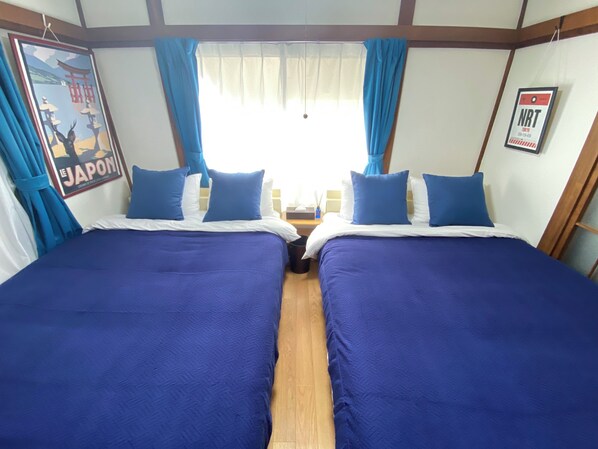 Habitación doble con dos camas, cada una con ropa de cama azul y almohadas blancas. Se observa una ventana con cortinas translúcidas y un cartel en la pared.