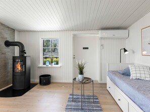 A cozy room with a wood-burning stove, a bed, a small table with a plant, and a window with a view of greenery.