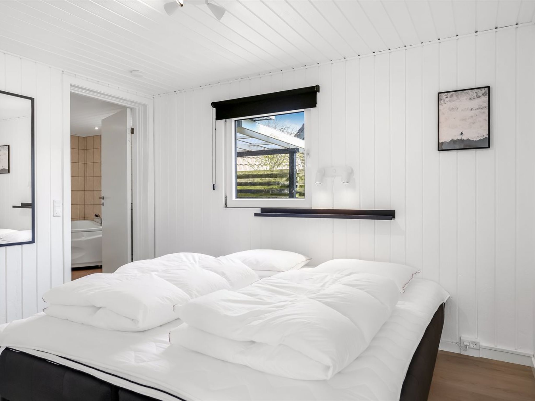 A bedroom with a bed, a window with a view, a mounted shelf, and a framed picture on the wall.