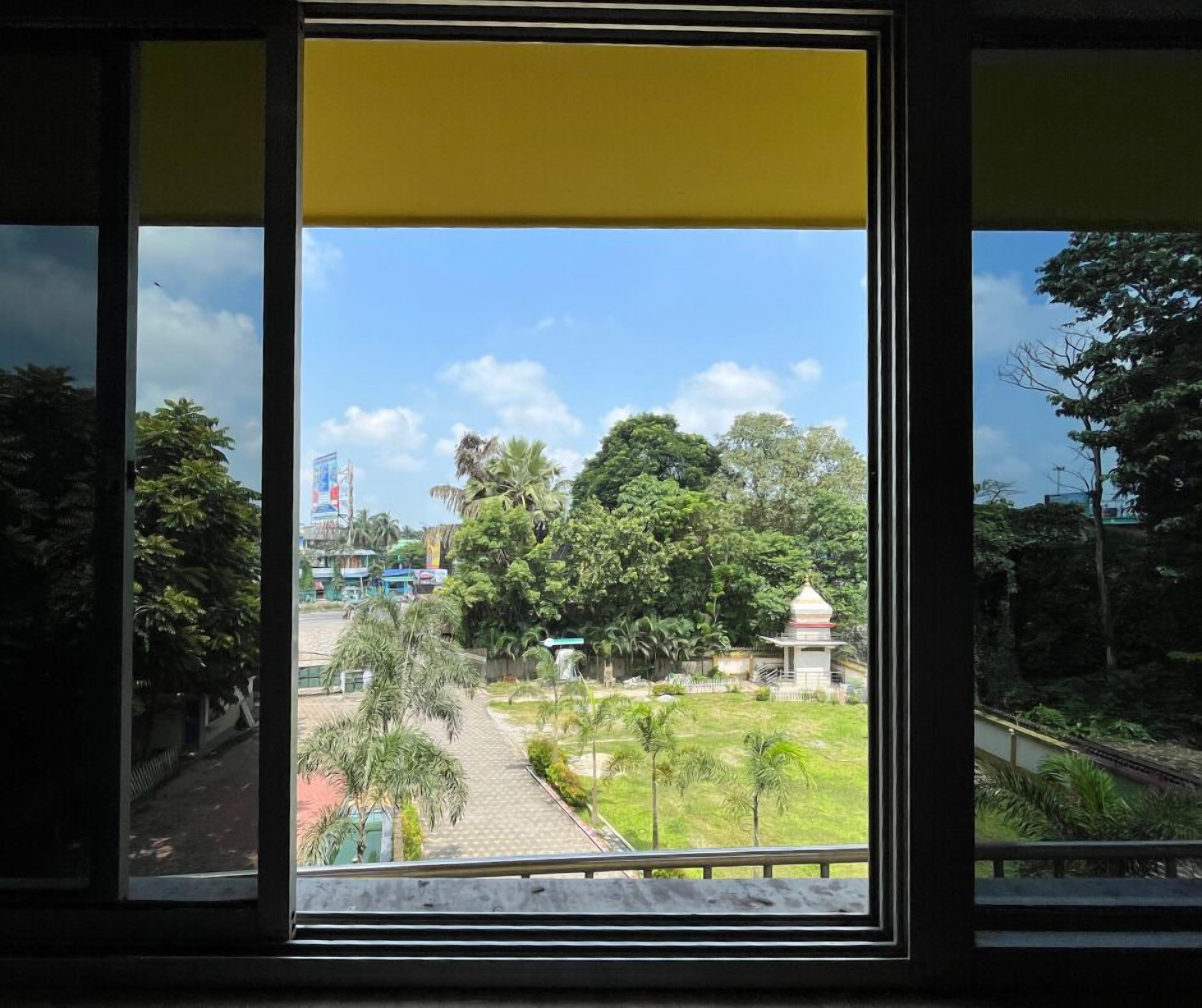 Vista a través de una ventana con una persiana amarilla, mostrando un camino que lleva a un templo rodeado de abundante vegetación.