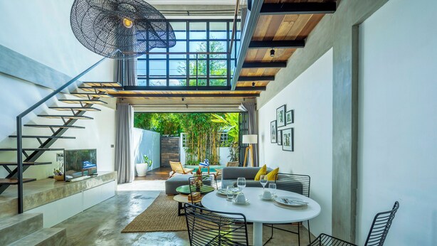 Un intérieur moderne avec un escalier menant à un balcon, un coin repas avec une table ronde et des chaises, et une vue sur la verdure à l’extérieur.