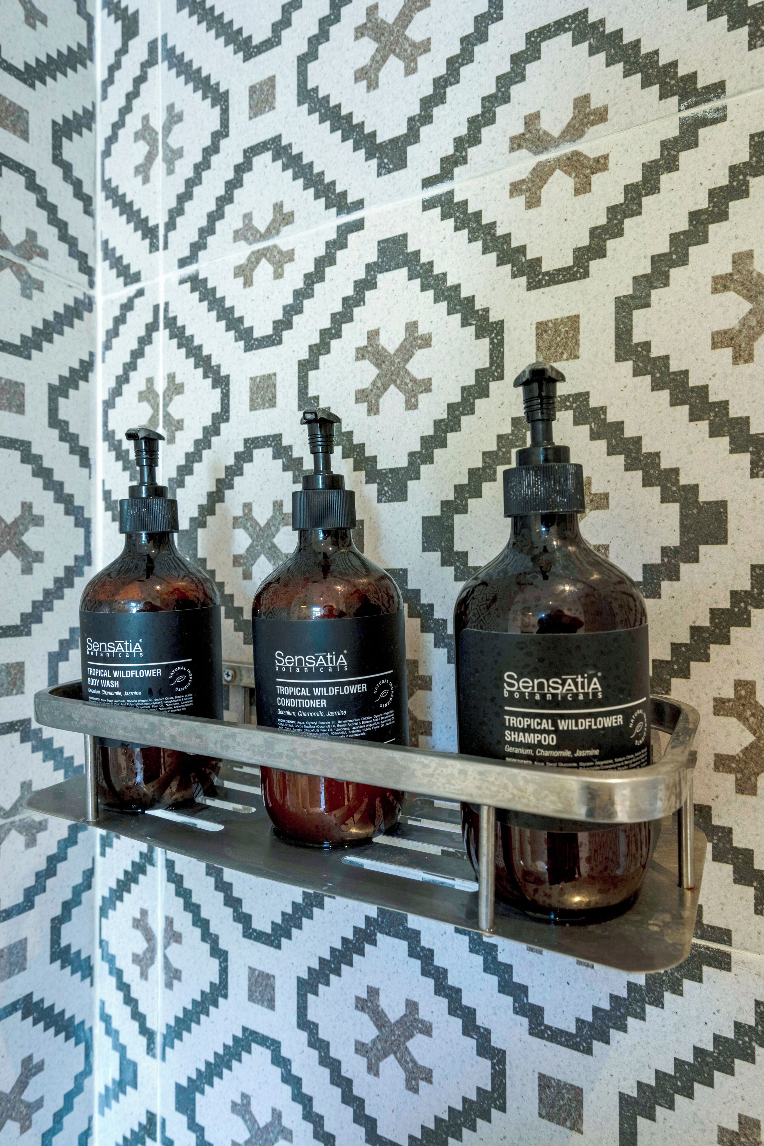 Three bottles of Sensatia Botanical shampoo on a metal shelf against a patterned wall.