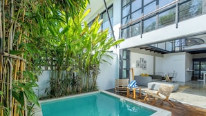 Un espace piscine intérieur moderne agrémenté de plantes en bambou, un coin salon avec des fauteuils et un vaste séjour doté d’un canapé et d’une table à manger.