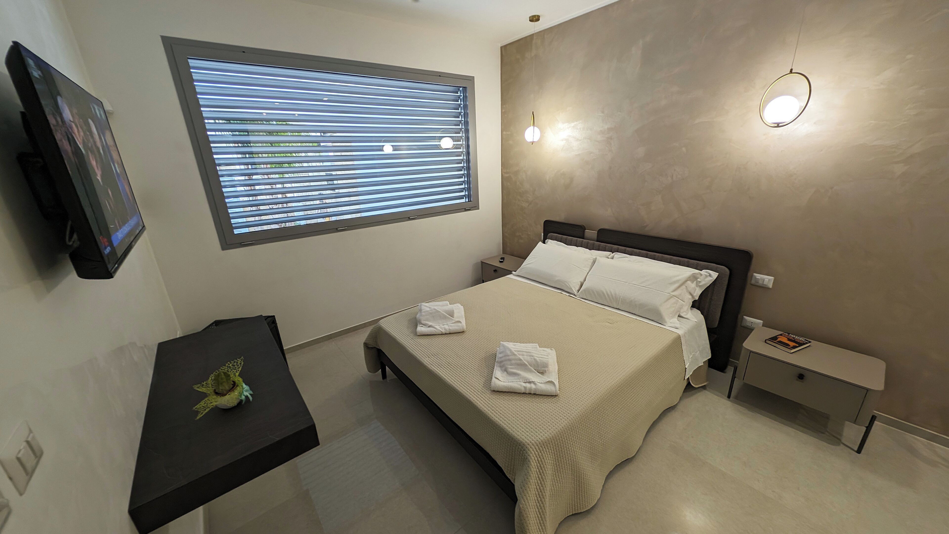 A modern hotel room with a bed, a small bedside table, a TV, and a window with blinds.