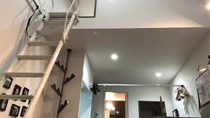 Un intérieur moderne avec un escalier blanc, des spots encastrés au plafond et un couloir menant à une pièce comprenant un lit et une table de chevet.