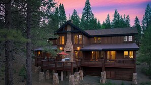 A large log cabin with multiple decks and a stone chimney, surrounded by tall pine trees.