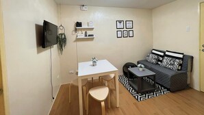 A compact living space with a TV mounted on the wall, a small table with a chair, a sofa with patterned cushions, and a ceiling light fixture.