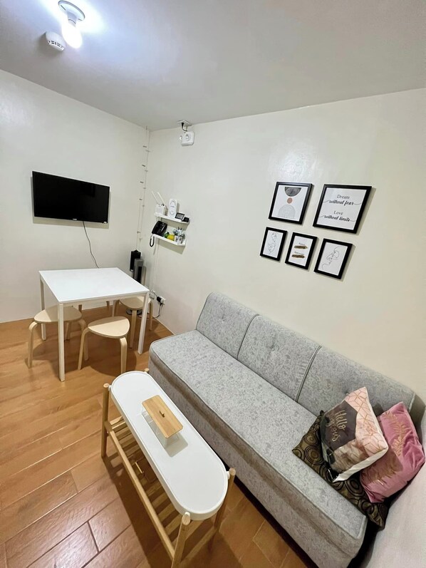 A living room with a grey sofa, a white table, and a TV mounted on the wall. There are framed pictures on the wall and a small shelf with a phone and other items. - Seawind Condo 2 BR Facing Samal View (Davao)