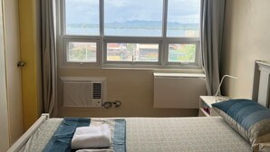 A bedroom with a bed, a bedside table, a window with a view of the city, and a radiator.