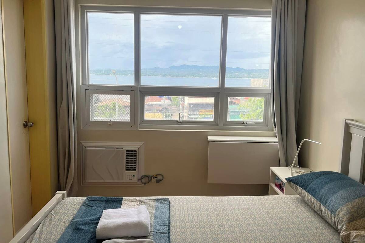 A bedroom with a bed, a bedside table, a window with a view of the city, and a radiator.