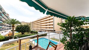 A balcony with a wooden table, a bottle of wine, and a chair. There is a pool in the background, surrounded by greenery and buildings. - Perfect Location 3 Bedrooms 2 bath, Parking, Pool REF048 (Fuengirola)