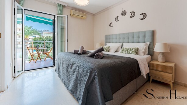 A bedroom with a bed, a nightstand, a lamp, a balcony with a table and chairs, and a view of buildings and greenery outside. - Perfect Location 3 Bedrooms 2 bath, Parking, Pool REF048 (Fuengirola)