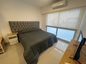 A modern bedroom with a tufted headboard, a large bed, a wooden nightstand, a flat-screen TV, and a large window with a view of buildings. - Modern Studio With Heated Pool in the Heart of San Telmo (Buenos Aires)
