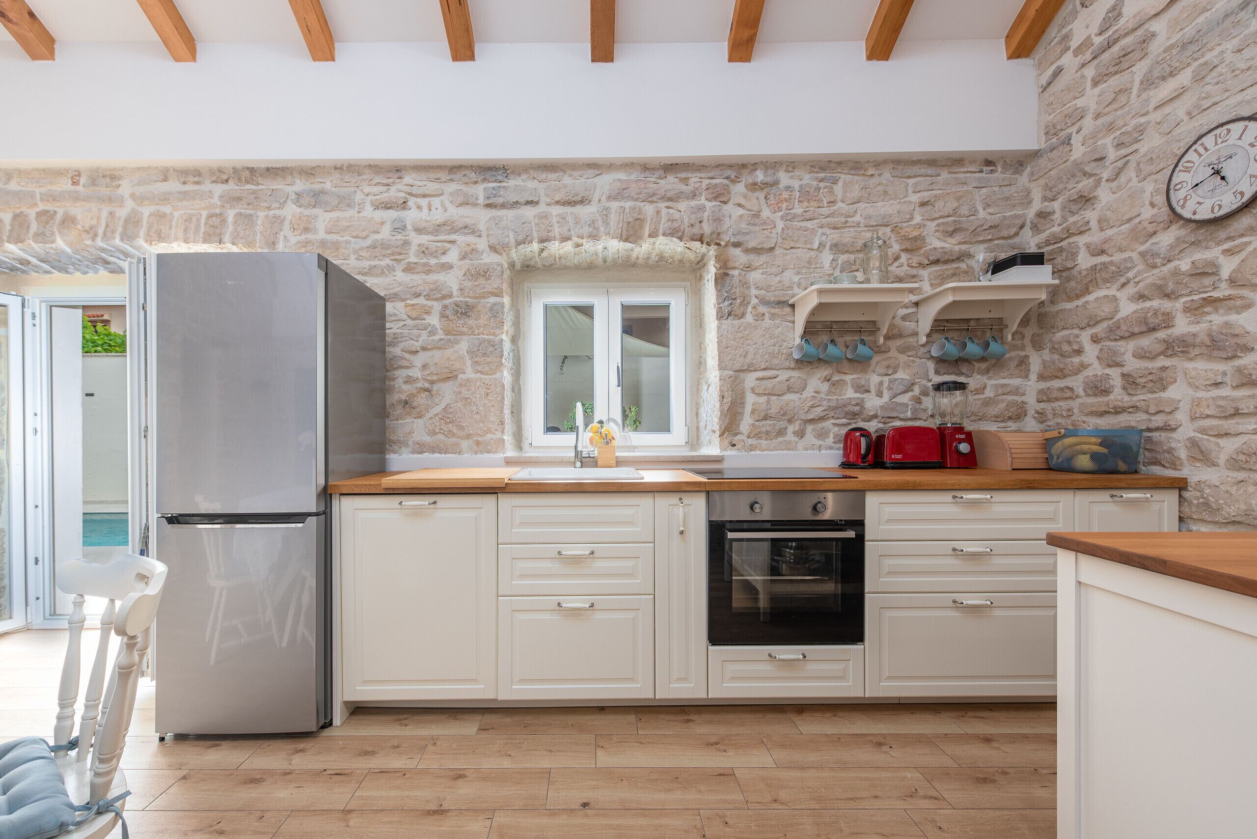 A kitchen with white cabinets, a stainless steel refrigerator, a wooden countertop, and a stone wall.