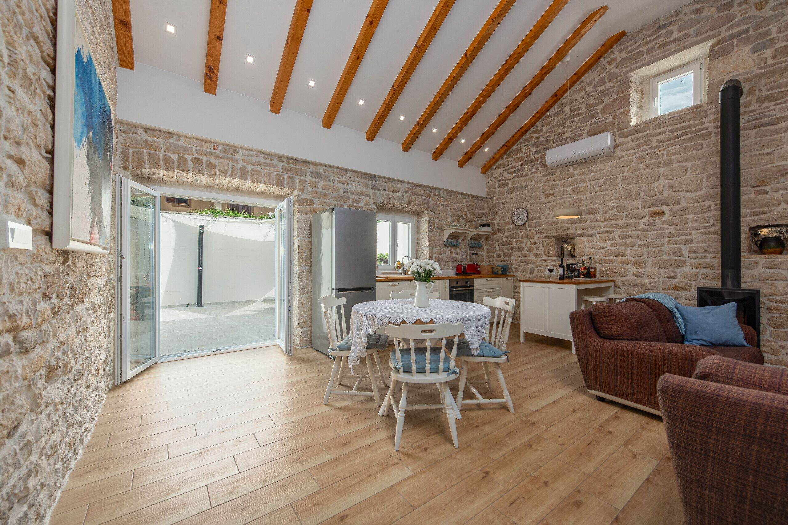 A spacious living area with a stone wall, wooden beams, a dining table with chairs, a sofa, and a kitchen area with a stove.