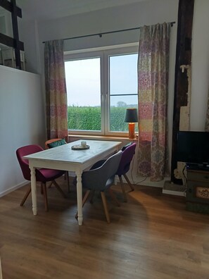A dining area with a table and chairs, a window with curtains, a lamp, and a television.