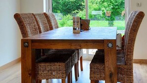 A dining area with a wooden table and wicker chairs, large windows with a view of greenery, and recessed ceiling lighting.