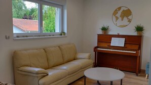 A room with a beige leather sofa, a wooden piano, a small table, and a window with a view of a red-roofed building.