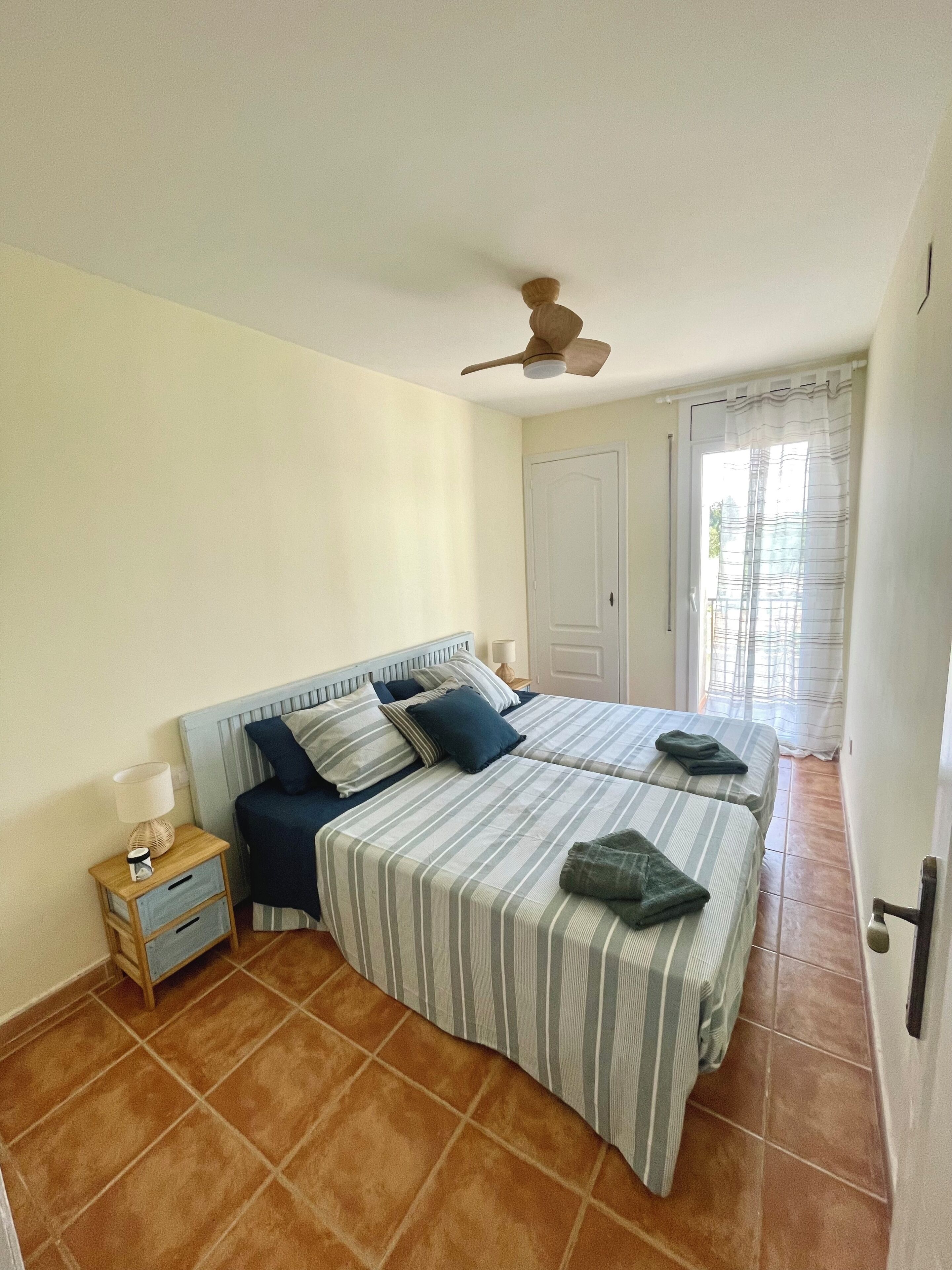 A bedroom with two beds, a nightstand, a ceiling fan, and a door leading to a balcony.