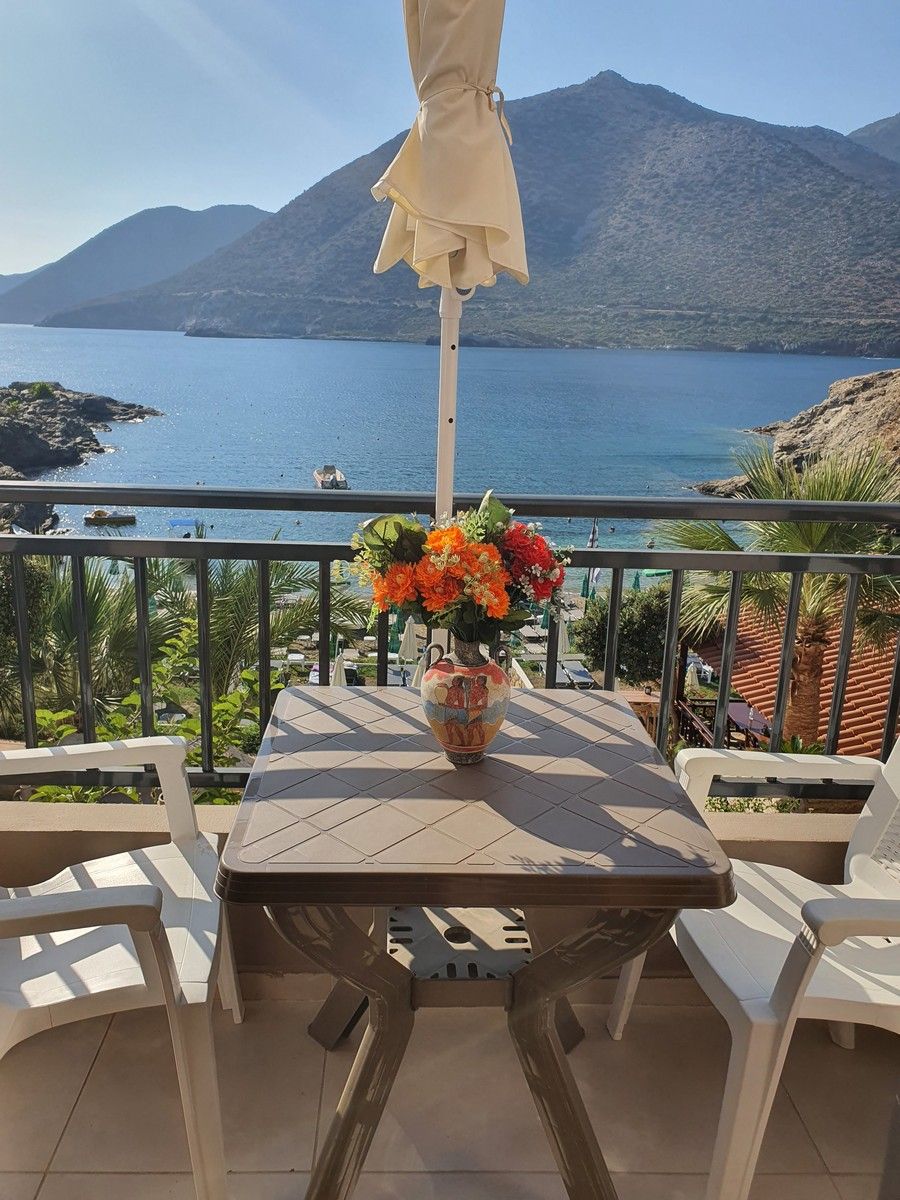 A balcony with a table, chairs, and a vase of flowers. A mountain and body of water are visible in the background.