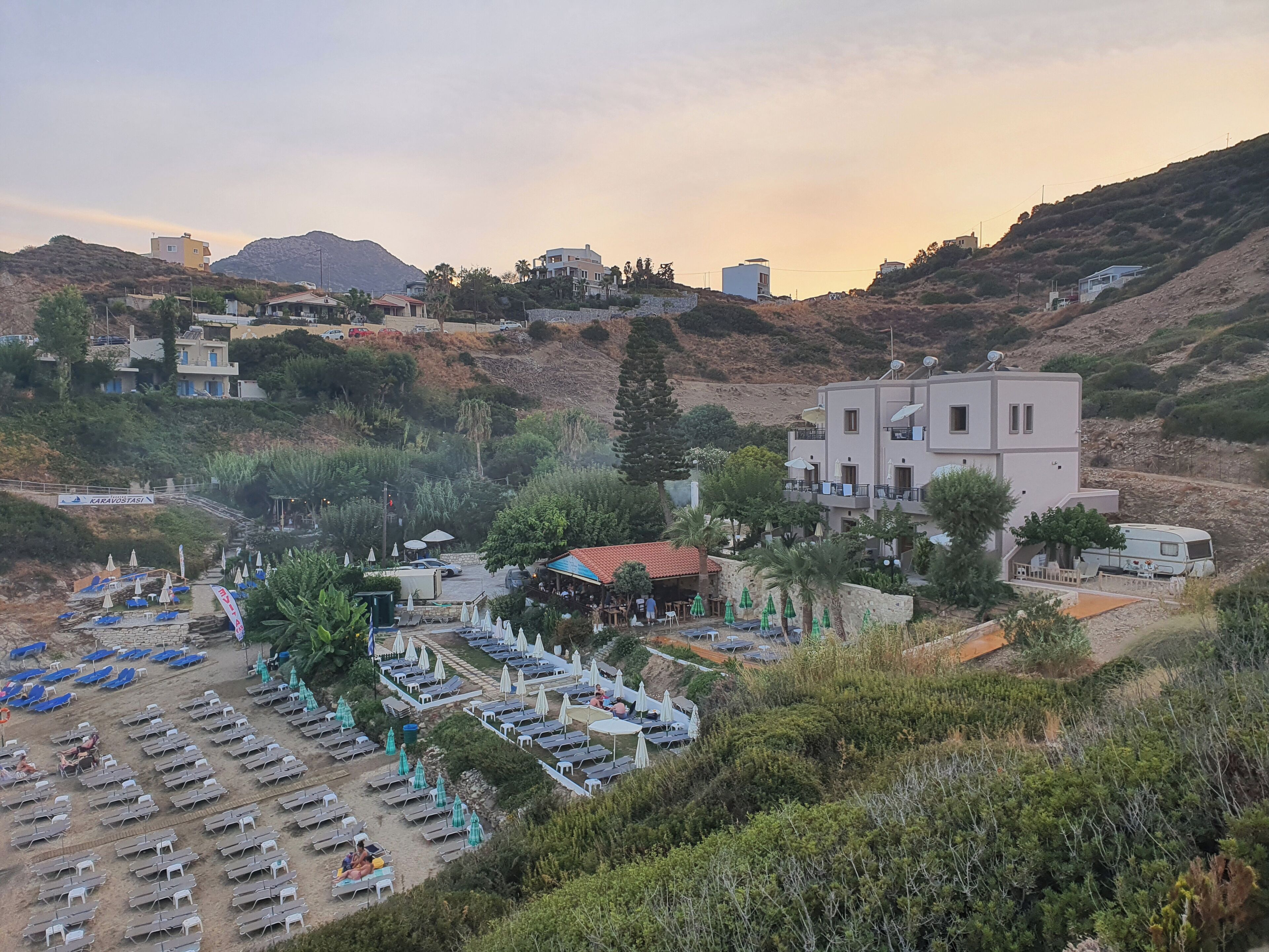 A coastal resort with multiple buildings, a beach area with sun loungers and umbrellas, and a mountainous backdrop.