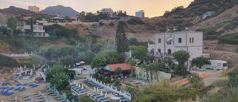 A coastal resort with multiple buildings, a beach area with sun loungers and umbrellas, and a mountainous backdrop.