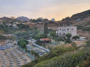 A coastal resort with multiple buildings, a beach area with sun loungers and umbrellas, and a mountainous backdrop. - Evita Studios & Apartments (Mylopotamos)
