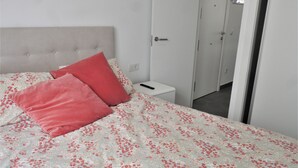 Un lit bien fait, recouvert d’une couvre-lit à motifs floraux et agrémenté de deux oreillers rouges, une table de chevet avec une télécommande, et une porte donnant sur une autre pièce.