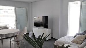 Un salon moderne avec un téléviseur à écran plat posé sur un meuble blanc, une table de salle à manger ronde entourée de chaises, un canapé avec des coussins et une grande plante en pot.