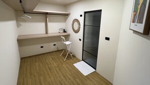 Een kleine kamer met een houten vloer, een witte stoel, een bureau met een kapstok en een deur, plus een ingelijst schilderij aan de muur.