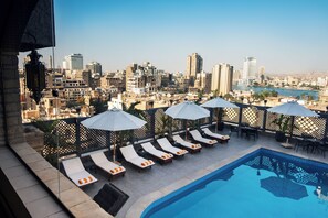 A rooftop pool area with lounge chairs and umbrellas, overlooking a cityscape with buildings and a river. - Marwa Palace Hotel (Giza)