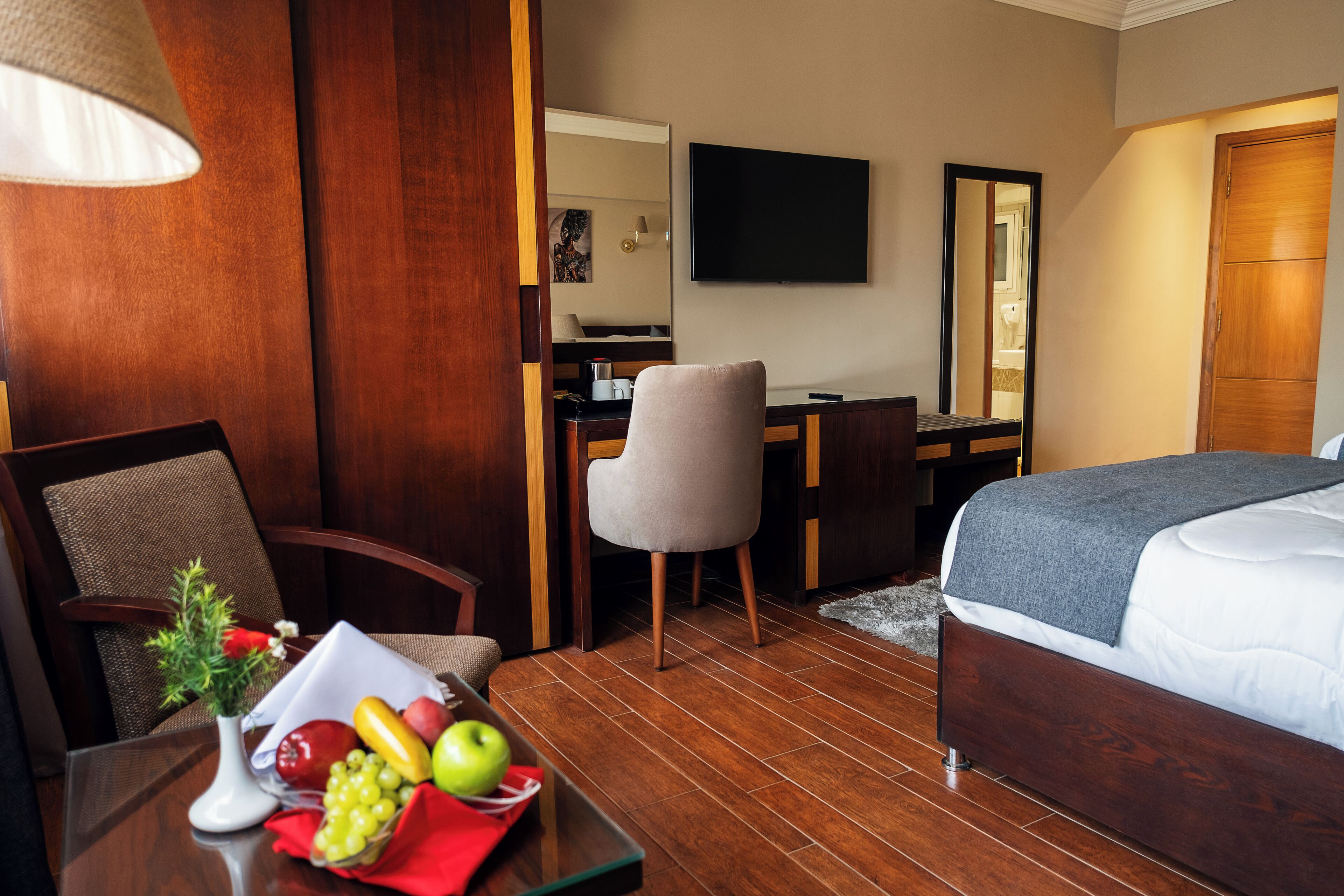 Quarto de hotel com piso de madeira, uma cama, uma escrivaninha com cadeira, uma televisão e uma fruteira na mesa.