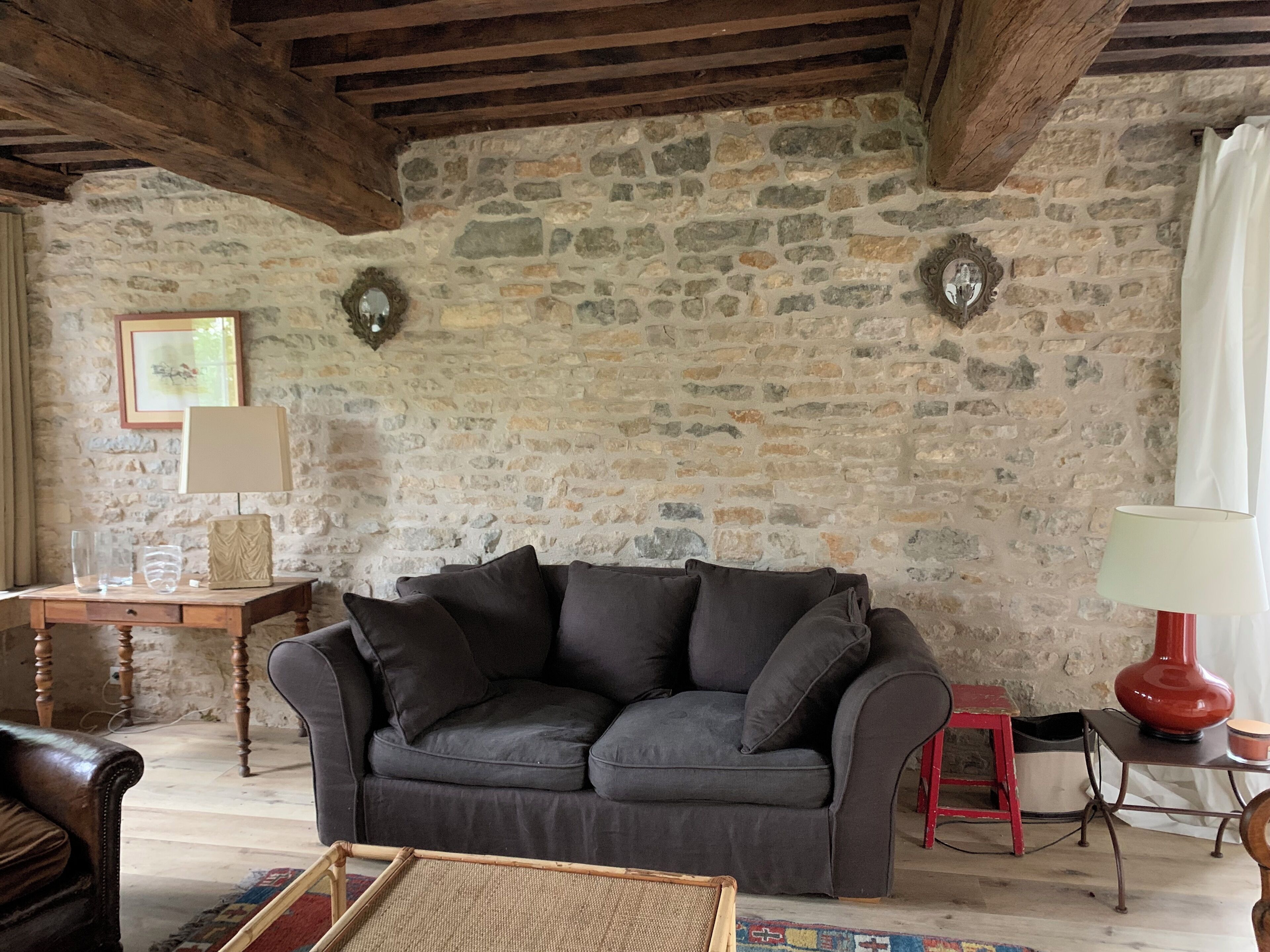 Una habitación rústica con pared de piedra, sofá gris oscuro, mesa de madera y un taburete rojo.