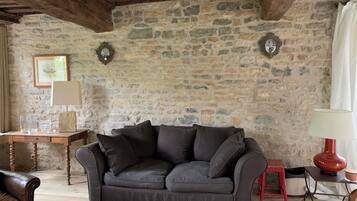 Una habitación rústica con pared de piedra, sofá gris oscuro, mesa de madera y un taburete rojo.