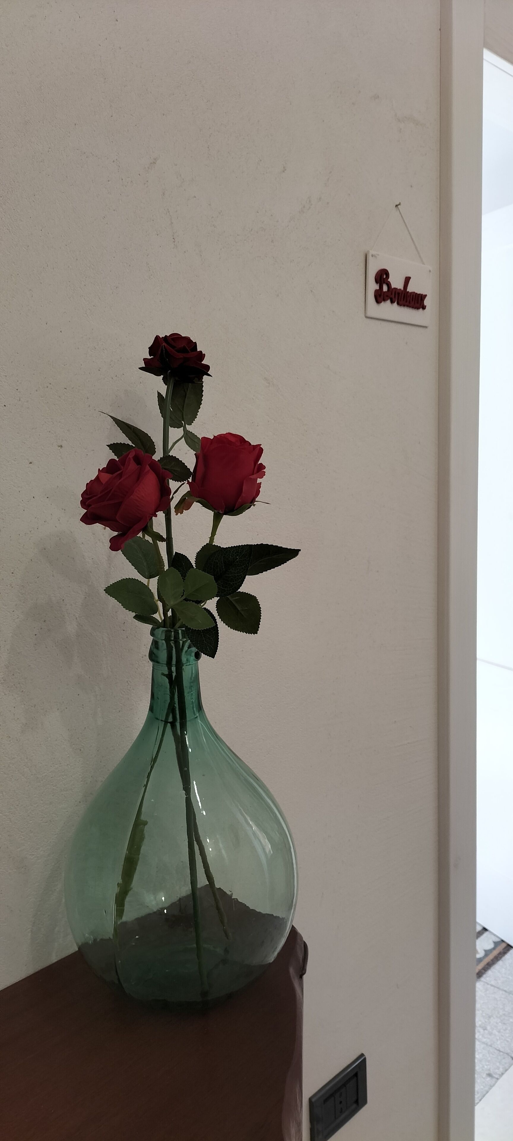 Un vase vert contenant trois roses rouges, posé sur une surface en bois devant un mur uni.