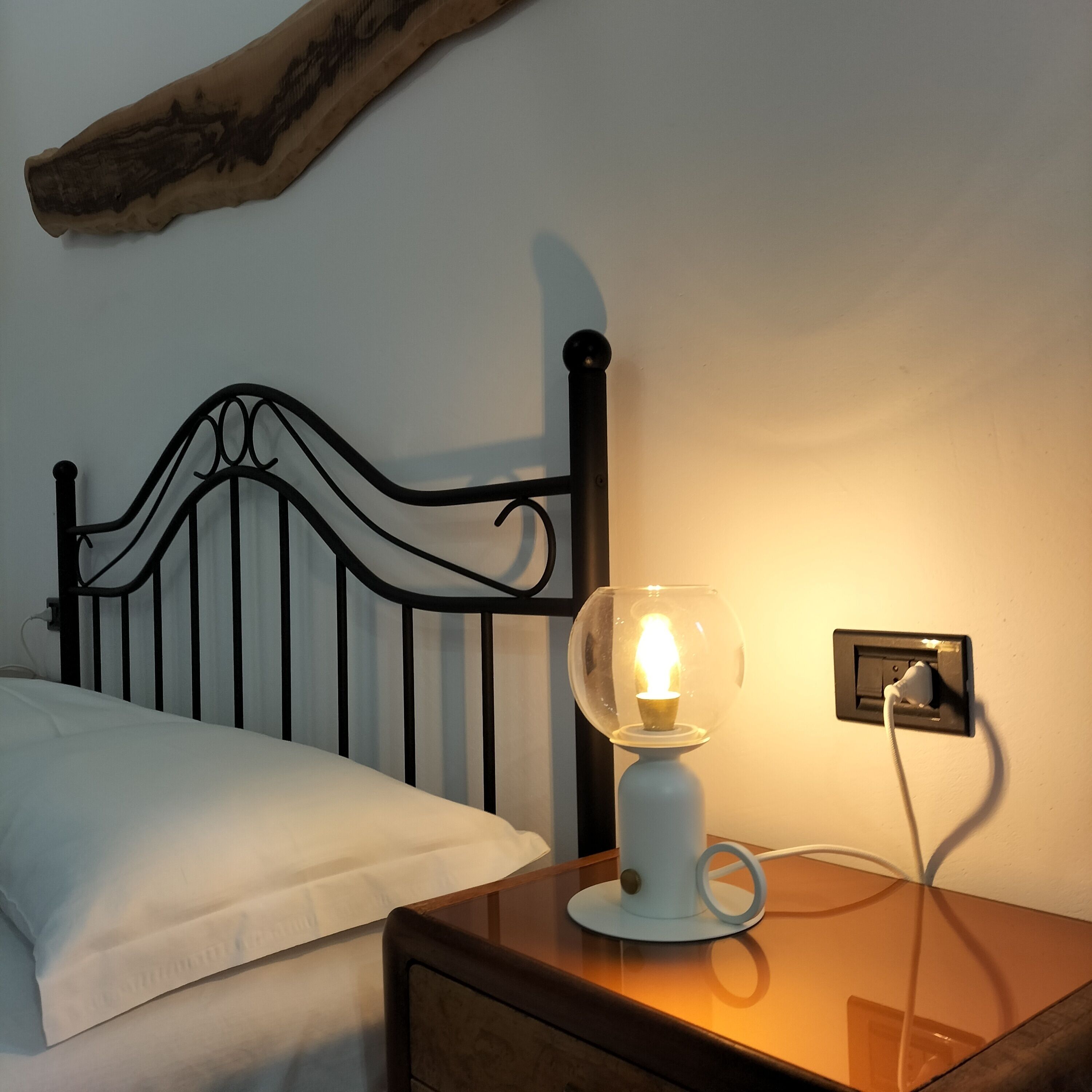Uma cama com roupa de cama branca, cabeceira de metal preto e mesa de cabeceira com abajur branco.