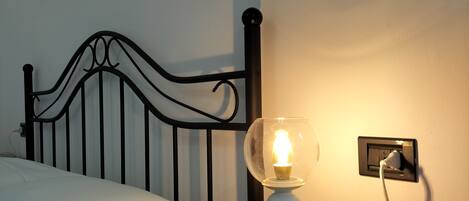 Uma cama com roupa de cama branca, cabeceira de metal preto e mesa de cabeceira com abajur branco.