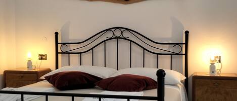 A bed with a metal frame, white bedding, and two red pillows. There are two bedside tables with lamps. A wooden piece hangs on the wall above the bed.
