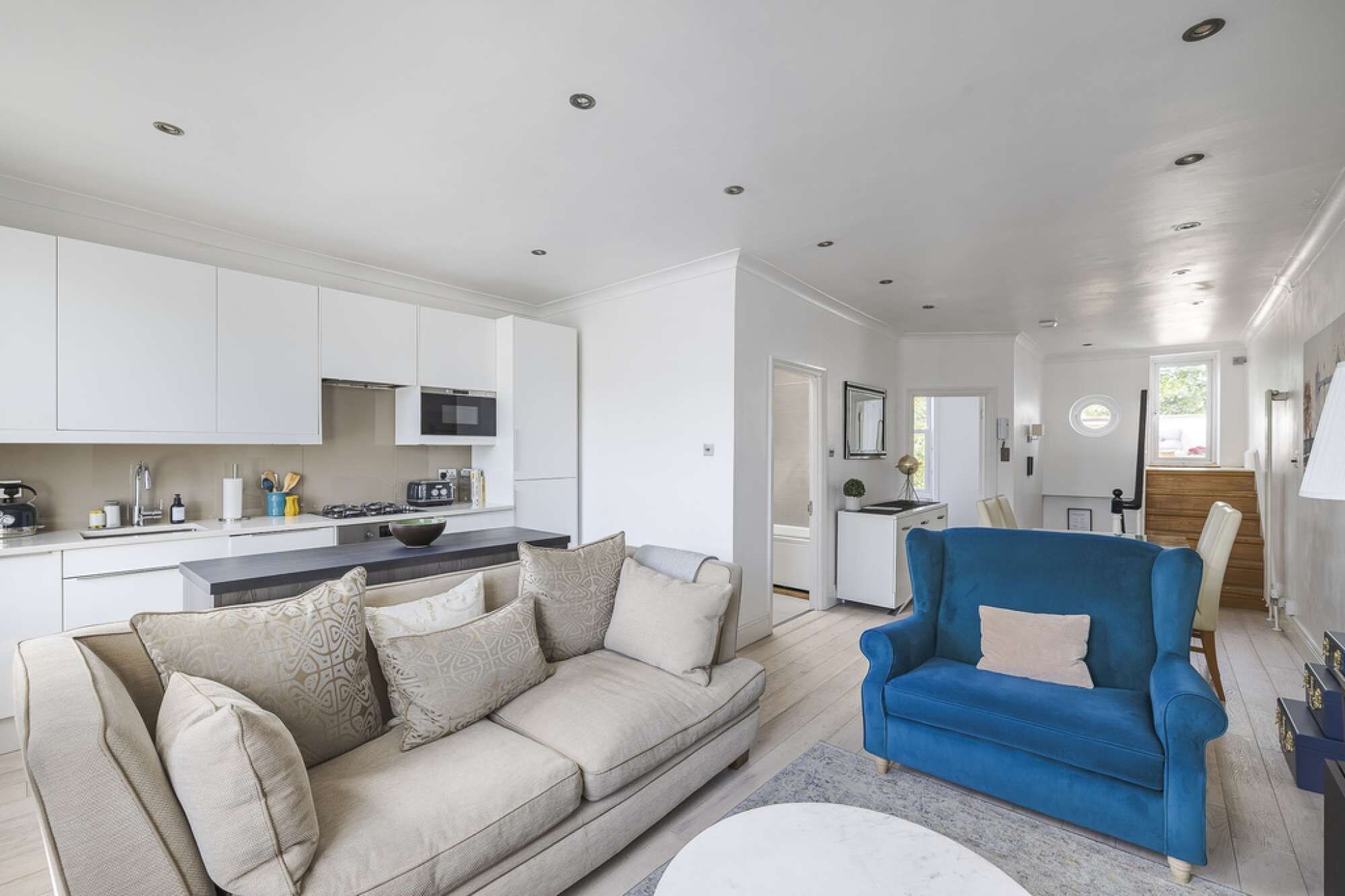 A modern living room with a beige sofa, a blue armchair, and a kitchen area with white cabinets and a microwave.